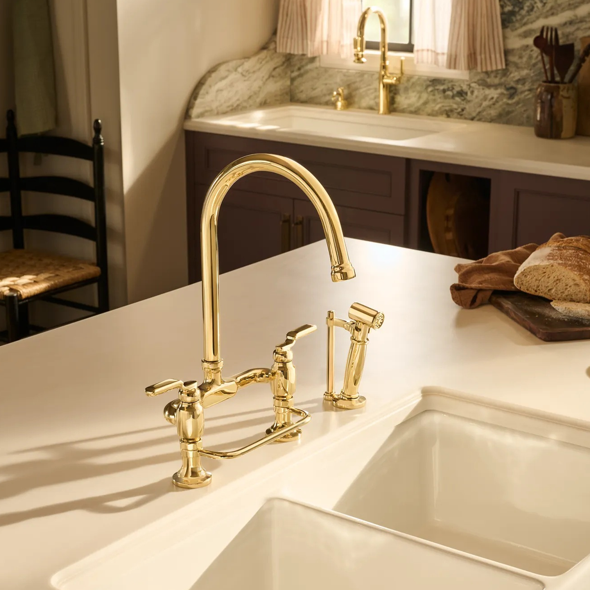 Brizo Faircroft deck-mount bridge faucet with arc spout and side spray in unlaquered brass installed on a white farmhouse sink countertop in a contemporary kitchen