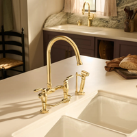 Brizo Faircroft deck-mount bridge faucet with arc spout and side spray in unlaquered brass installed on a white farmhouse sink countertop in a contemporary kitchen