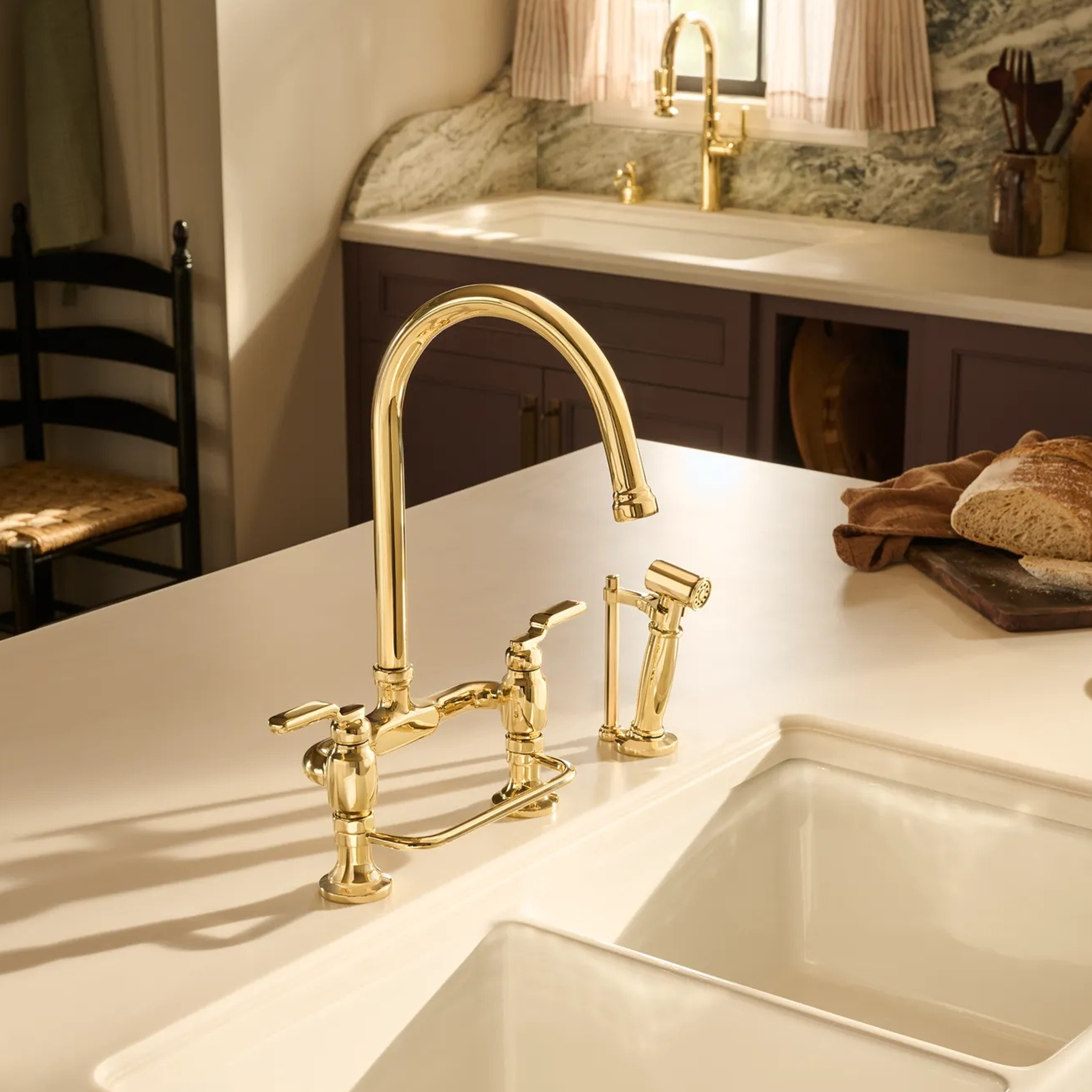 Brizo Faircroft deck-mount bridge faucet with arc spout and side spray in unlaquered brass installed on a white farmhouse sink countertop in a contemporary kitchen