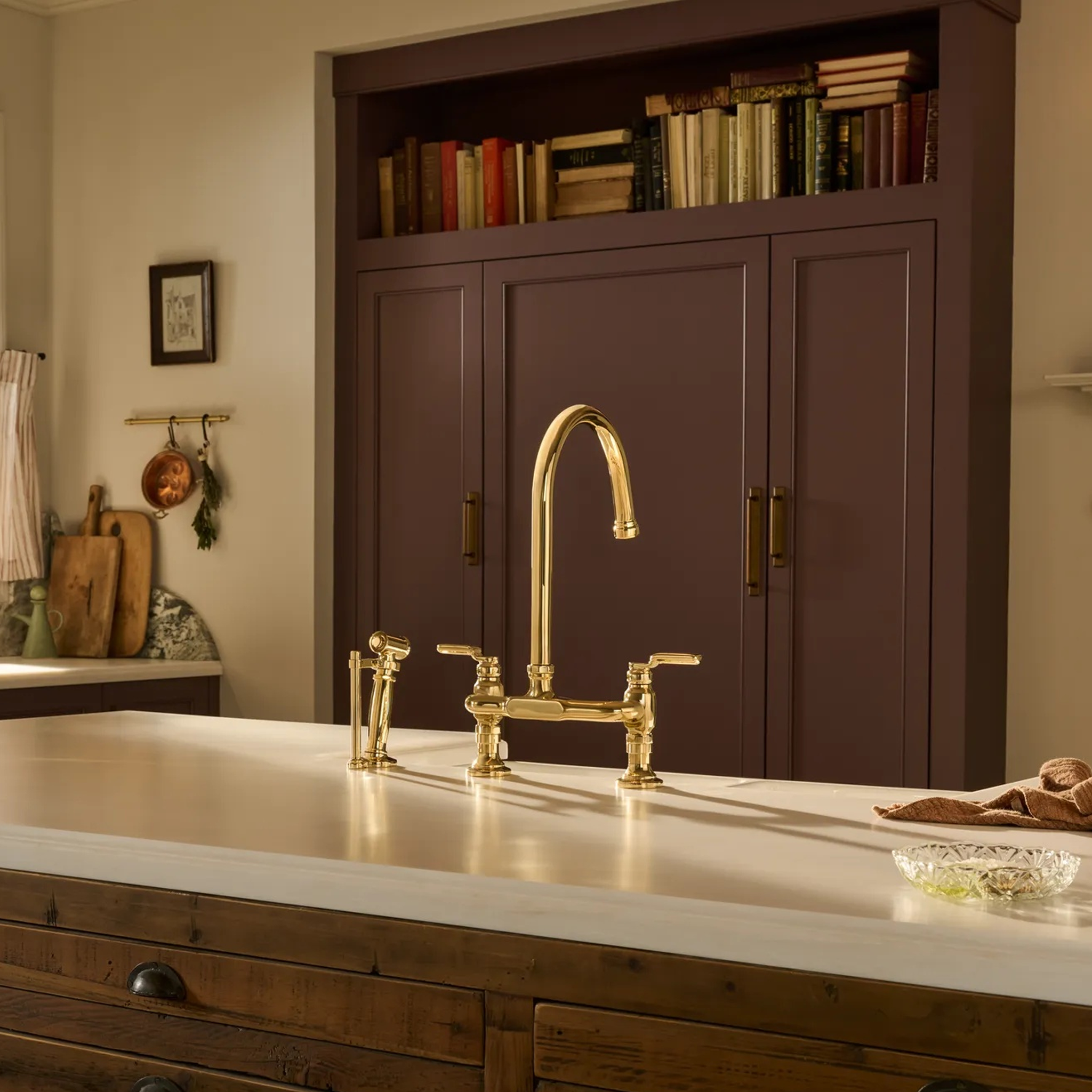 Brizo Faircroft deck-mount bridge faucet with arc spout and side spray in unlacquered brass finishe centered on a white kitchen island countertop.