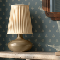 A Clementine table lamp with a pleated wheat-linen shade and a ceramic base, styled on a table against a patterned blue-green wallpaper background.