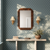 A rectangular wooden wall mirror with a dark brown frame hangs on a blue patterned wallpapered wall, flanked by two Eunice wall sconces above a rustic console table with a vase of dried flowers.