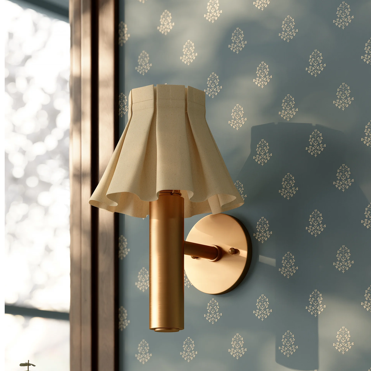 A close-up shot of modern Eunice wall sconce with a pleated cream fabric shade, mounted on a blue patterned wallpaper wall next to a window.