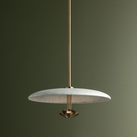 A Garth pendant light with a wide, shallow round white shade, a brushed brass suspension rod and geometric metal finial, shown against a dark olive green background.