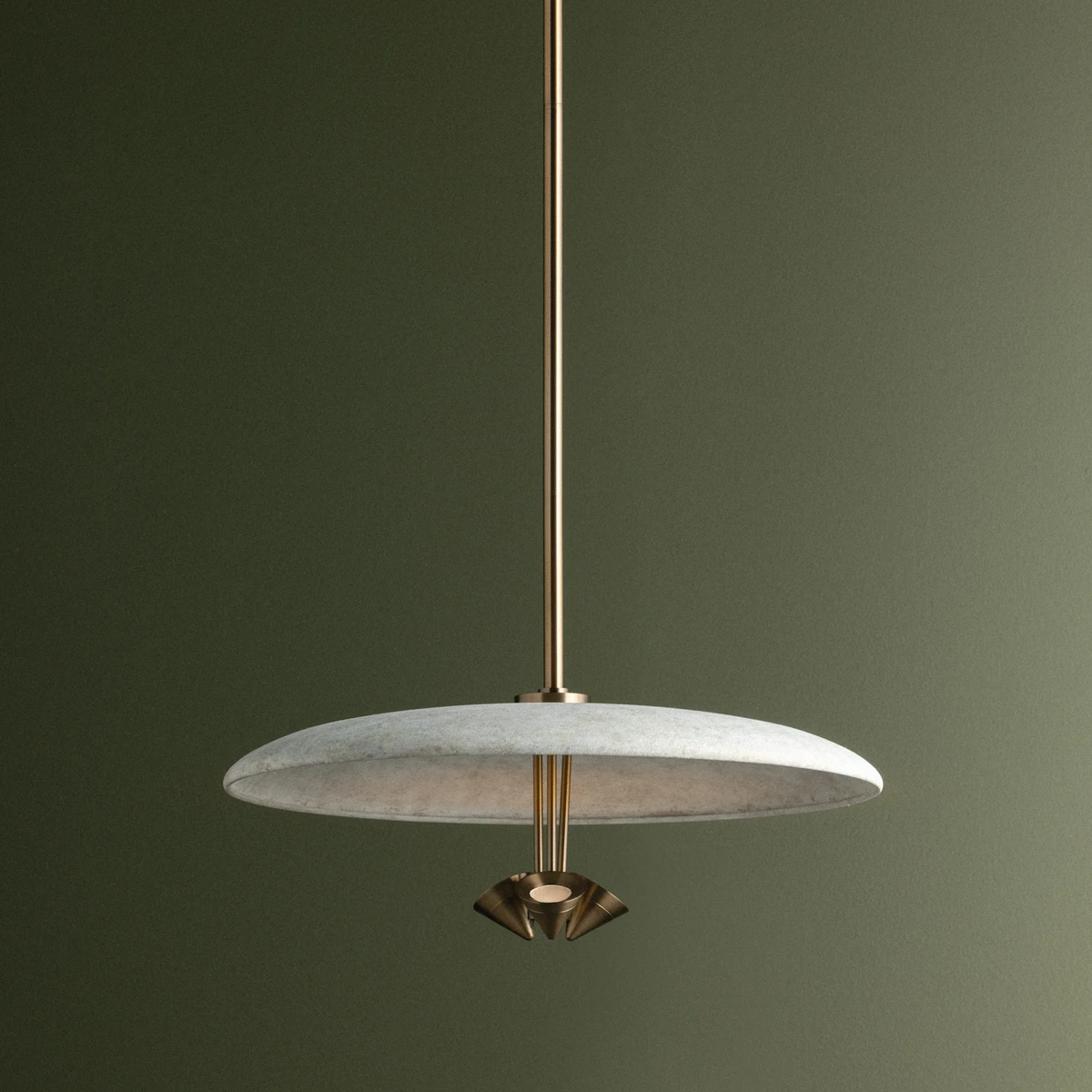 A Garth pendant light with a wide, shallow round white shade, a brushed brass suspension rod and geometric metal finial, shown against a dark olive green background.