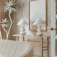 Two sculptural Kamas Table Lamps with tapered white shades sit on a rustic wood console table beneath a large framed artwork.