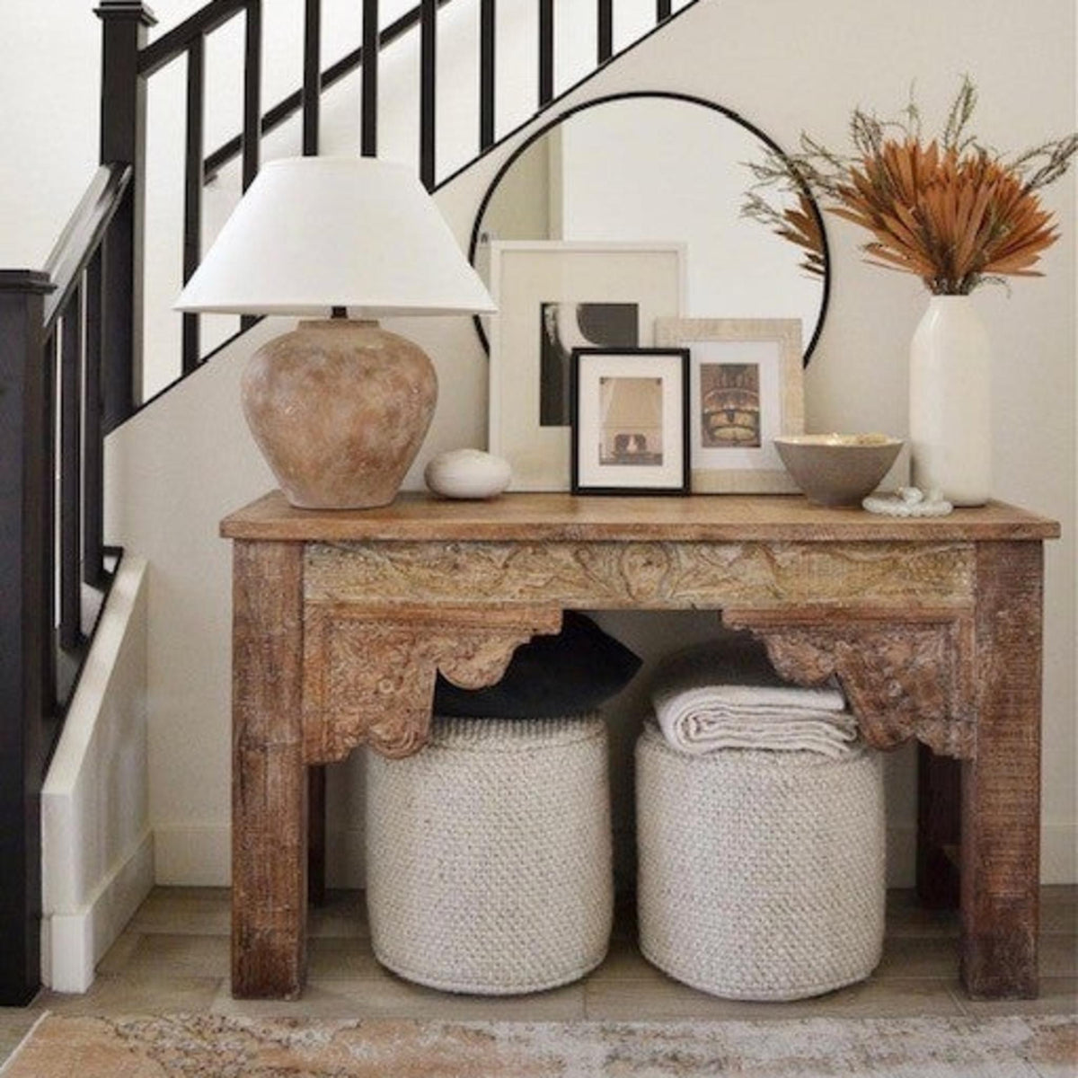 A rustic carved wooden console table styled in a home entryway next to a staircase, holding a Calabria Table Lamp, picture frames, a vase with dried grass.