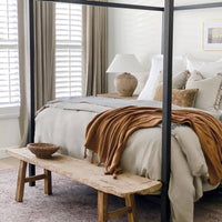 A styled bedroom scene featuring a four-poster frame bed with neutral beige bedding, a rust-brown throw blanket, and a Calabria Table Lamp