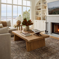 A rustic light brown wooden coffee table in a bright, modern living room with a fireplace, built-in shelves, and a Calabria Table Lamp