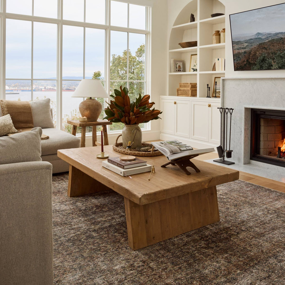 A rustic light brown wooden coffee table in a bright, modern living room with a fireplace, built-in shelves, and a Calabria Table Lamp