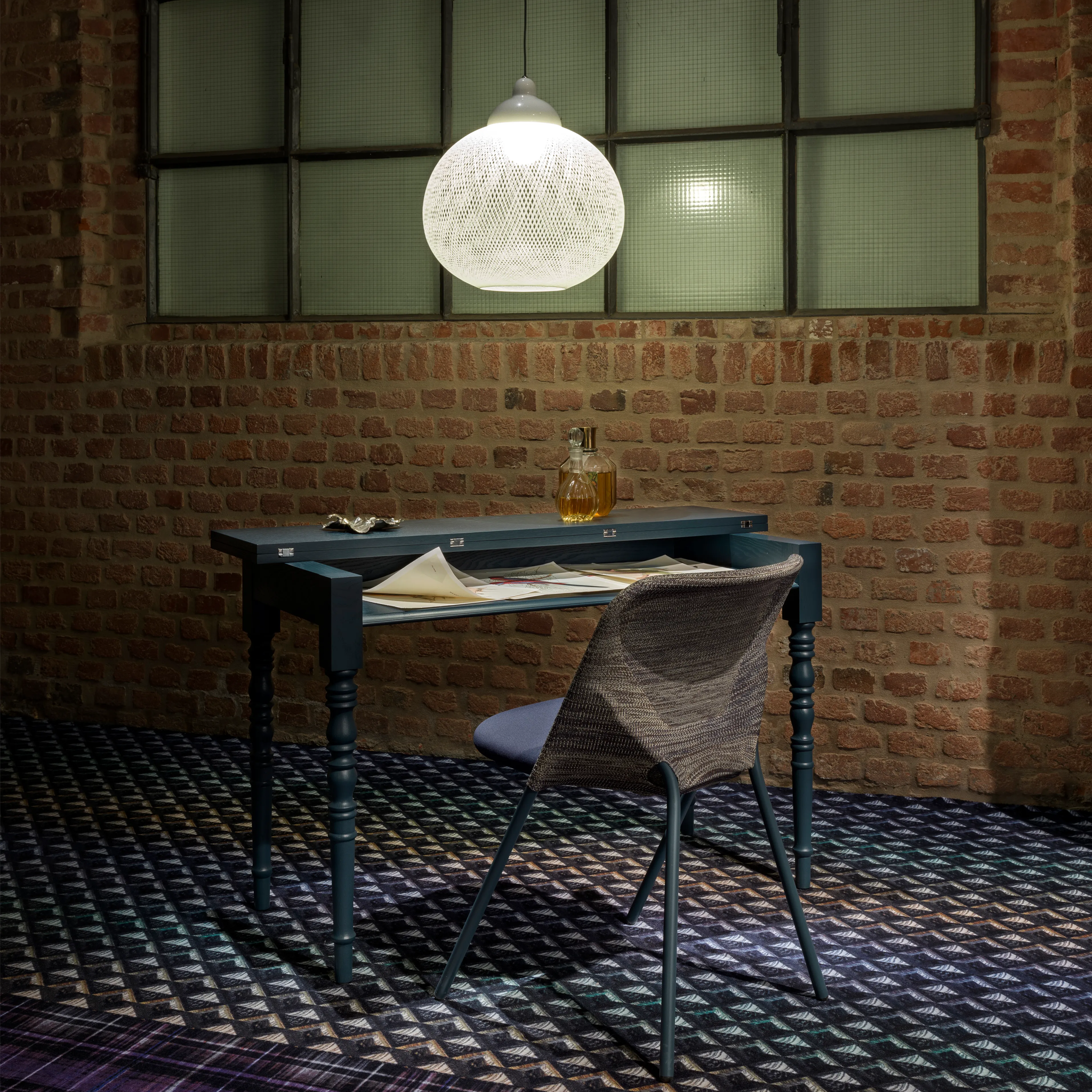 Desk with chair under a white Non Random light fixture in a room with brick walls and patterned carpet.