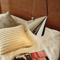 A modern cone-shaped Pose pendant lamp with a reflective silver metallic finish, hanging over a bed with cream bedding, textured pillows, and a brown woven headboard.
