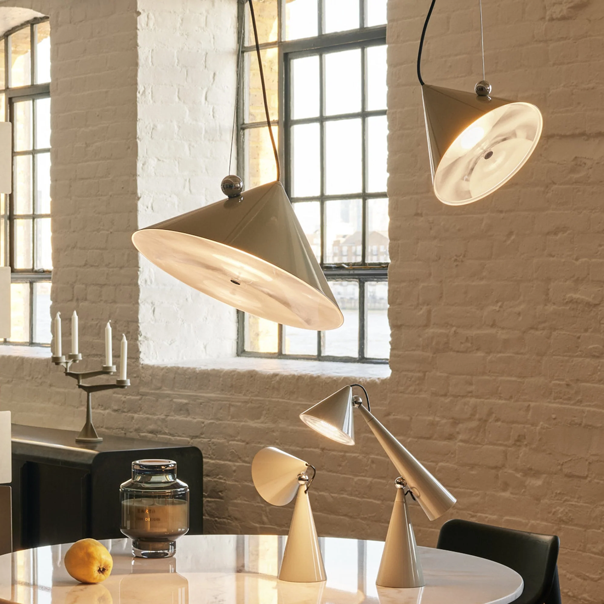 A modern dining room scene featuring a Pose collection from Tom Dixon. Set of pendants and table lamps in a warm cream and silver finish, styled on a white marble table against a white brick wall.
